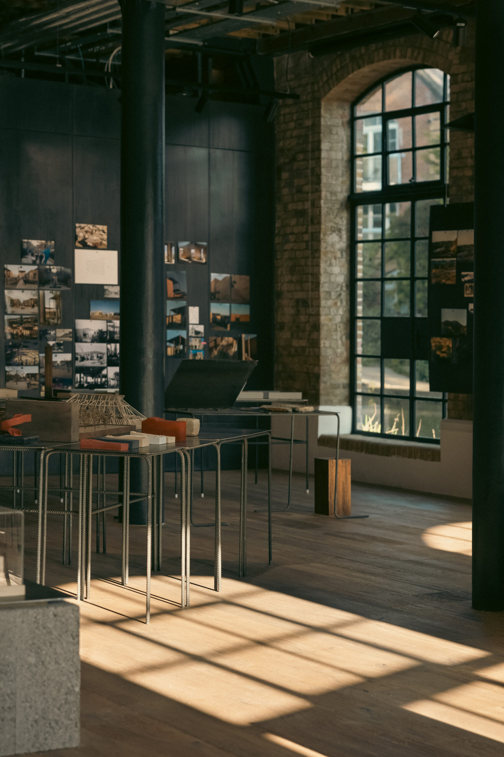 Detail view of Memory as Material at obra, London, showing architectural models and material samples on rebar-framed metal tables, with archival project photographs pinned to dark-painted walls, cast-iron columns and a steel-framed arched window casting geometric shadows across the oak floor.