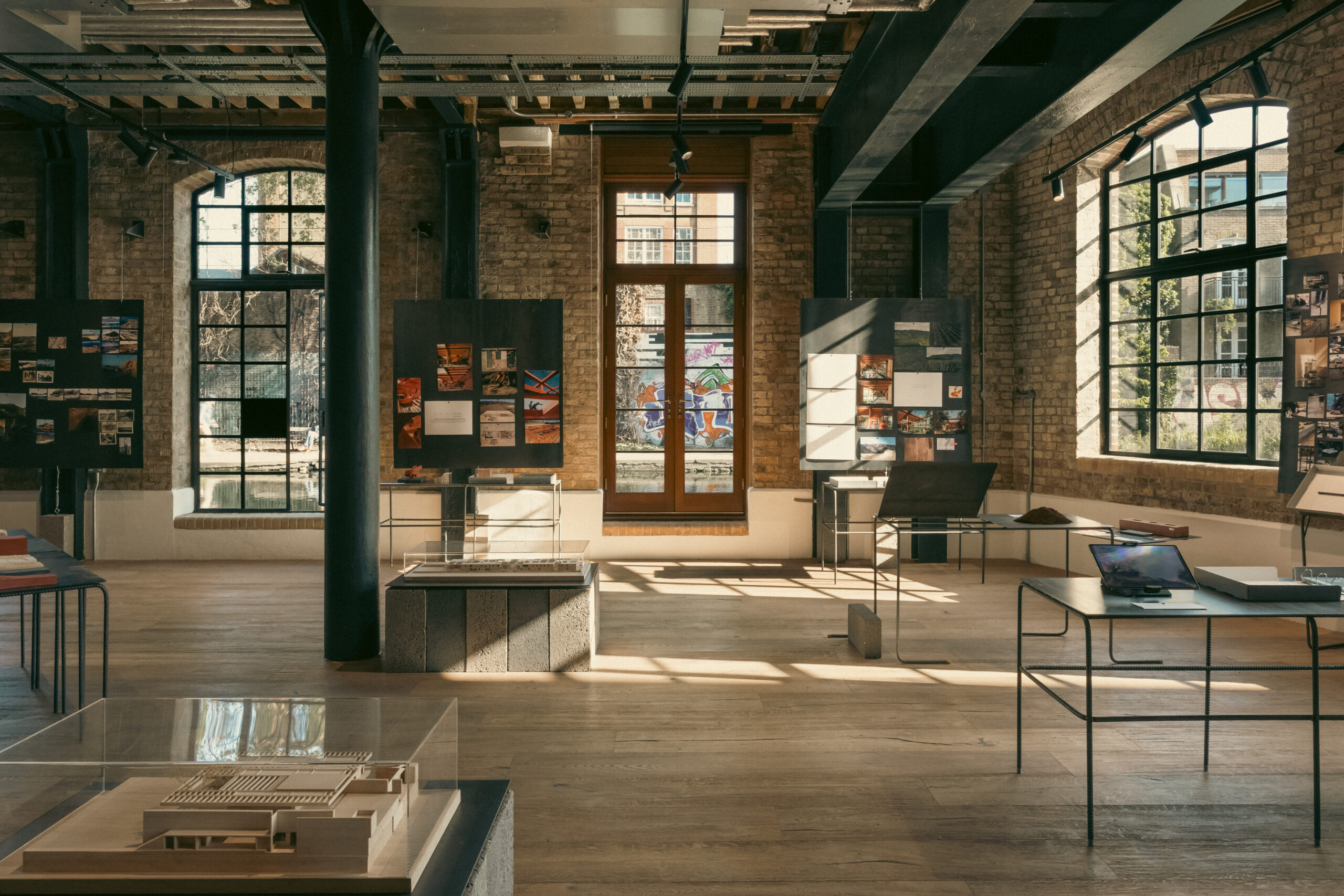 Sunlit interior view of the obra exhibition by Sordo Madaleno, with architectural scale models in glass cases, project photographs pinned to freestanding boards, and material samples on metal stands, set within a converted industrial space with exposed brick walls, cast-iron columns and large steel-framed windows overlooking a canal.