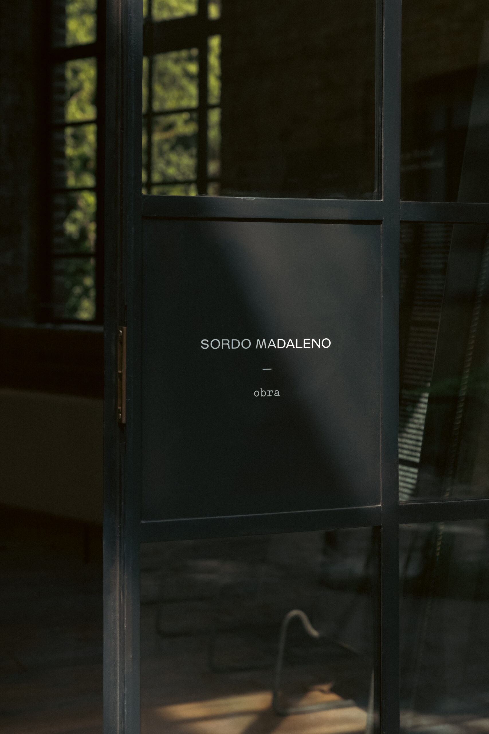 Close-up of a steel-framed glass door bearing the text "SORDO MADALENO — obra" in white lettering, with a garden visible through the surrounding windows.