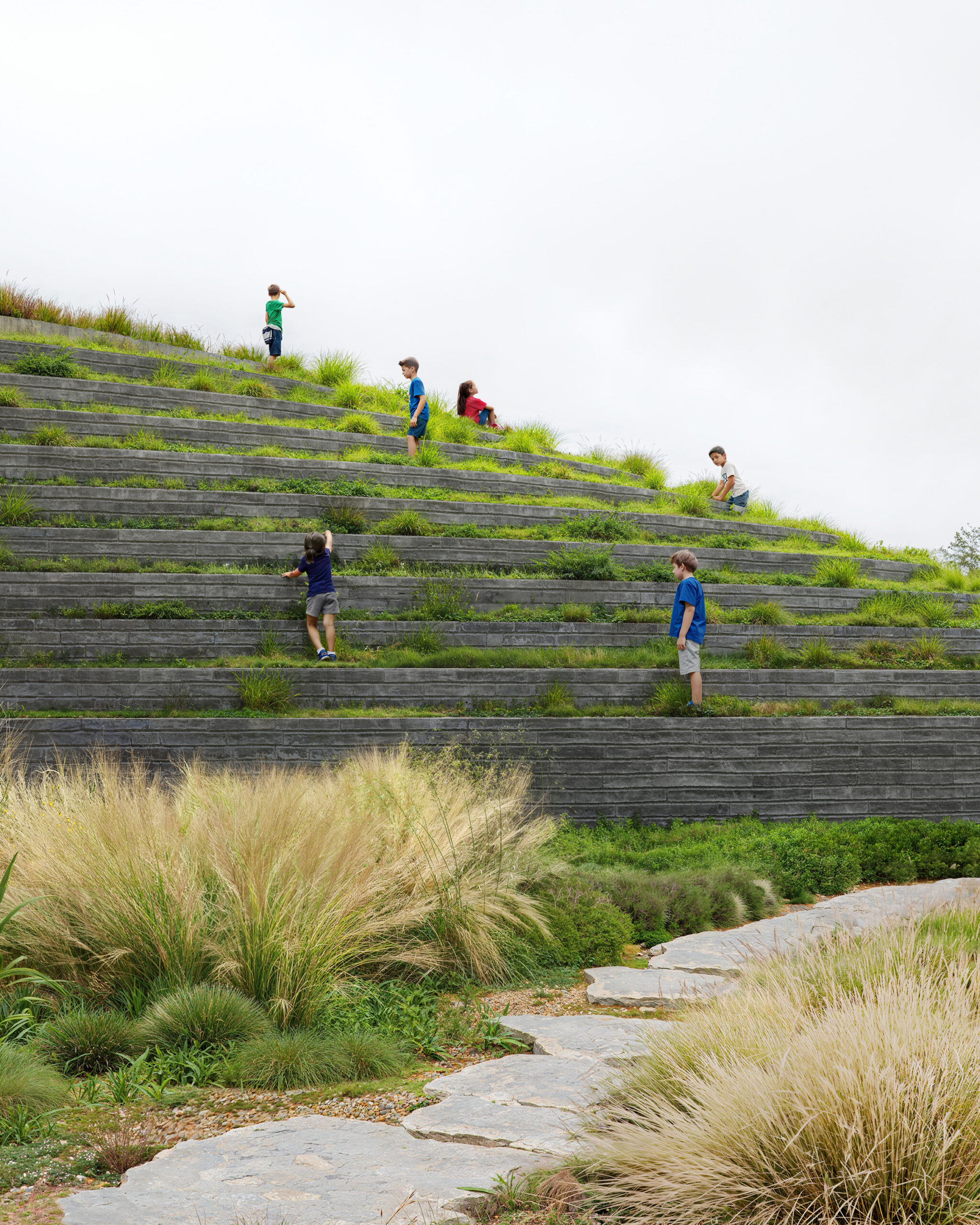 Children climbing grass-covered stepped terraces at Canadian School on volcanic terrain, Cholula, Puebla. Contemporary educational architecture in Mexico by Sordo Madaleno.