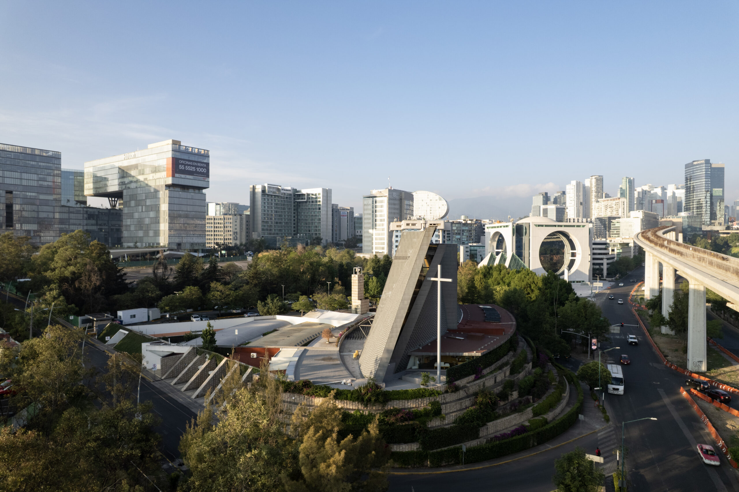 Vista aérea de la iglesia de San Josemaría Escrivá, con su forma triangular revestida de zinc, en Santa Fe, con el perfil de la Ciudad de México al fondo. Por Sordo Madaleno.