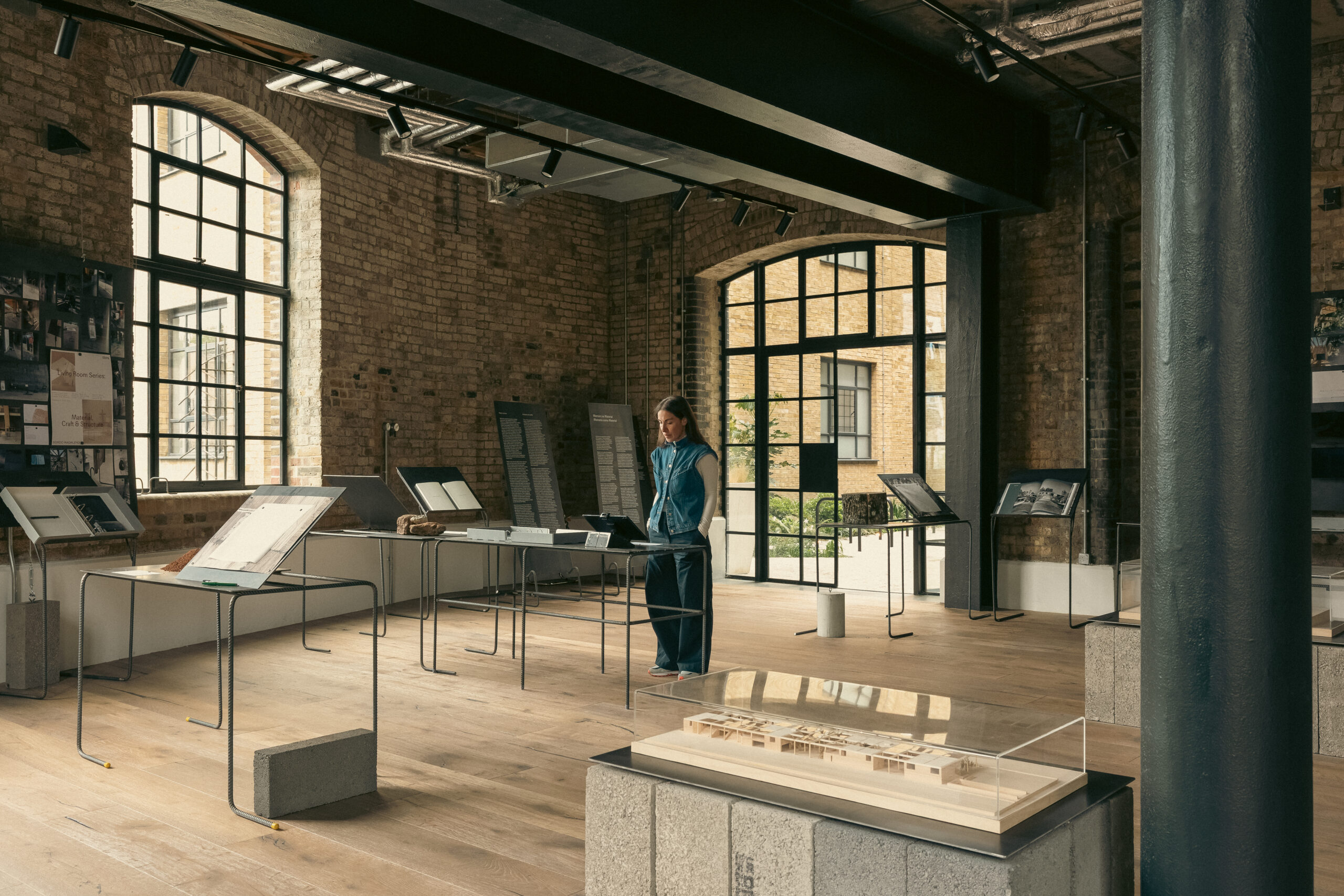 Interior view of the Memory as Material exhibition, showing a visitor examining architectural drawings and models displayed on slender metal stands within a converted industrial space with exposed brick walls and steel-framed windows.
