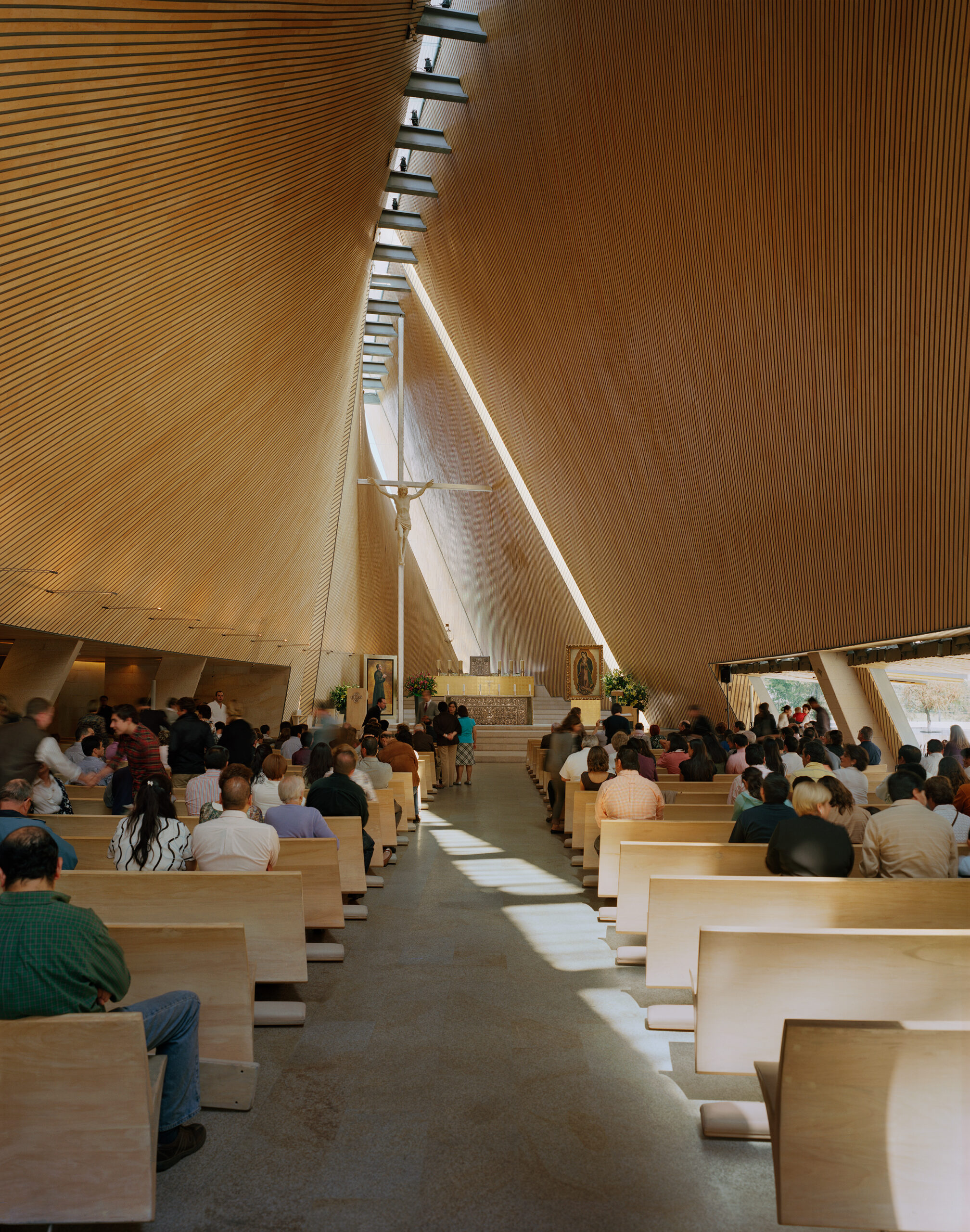 Nave interior de la iglesia de San Josemaría Escrivá durante la misa, con paredes curvas revestidas de madera y la Cruz de Luz sobre el altar. Obra de Sordo Madaleno.