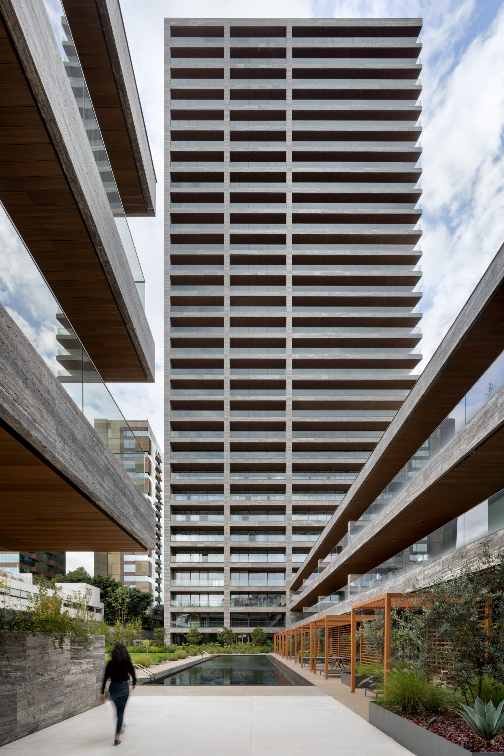 Ground-level exterior view of Rubén Darío 255's stone-clad tower by Sordo Madaleno, framed by a landscaped corridor with reflecting pool and timber pergola.