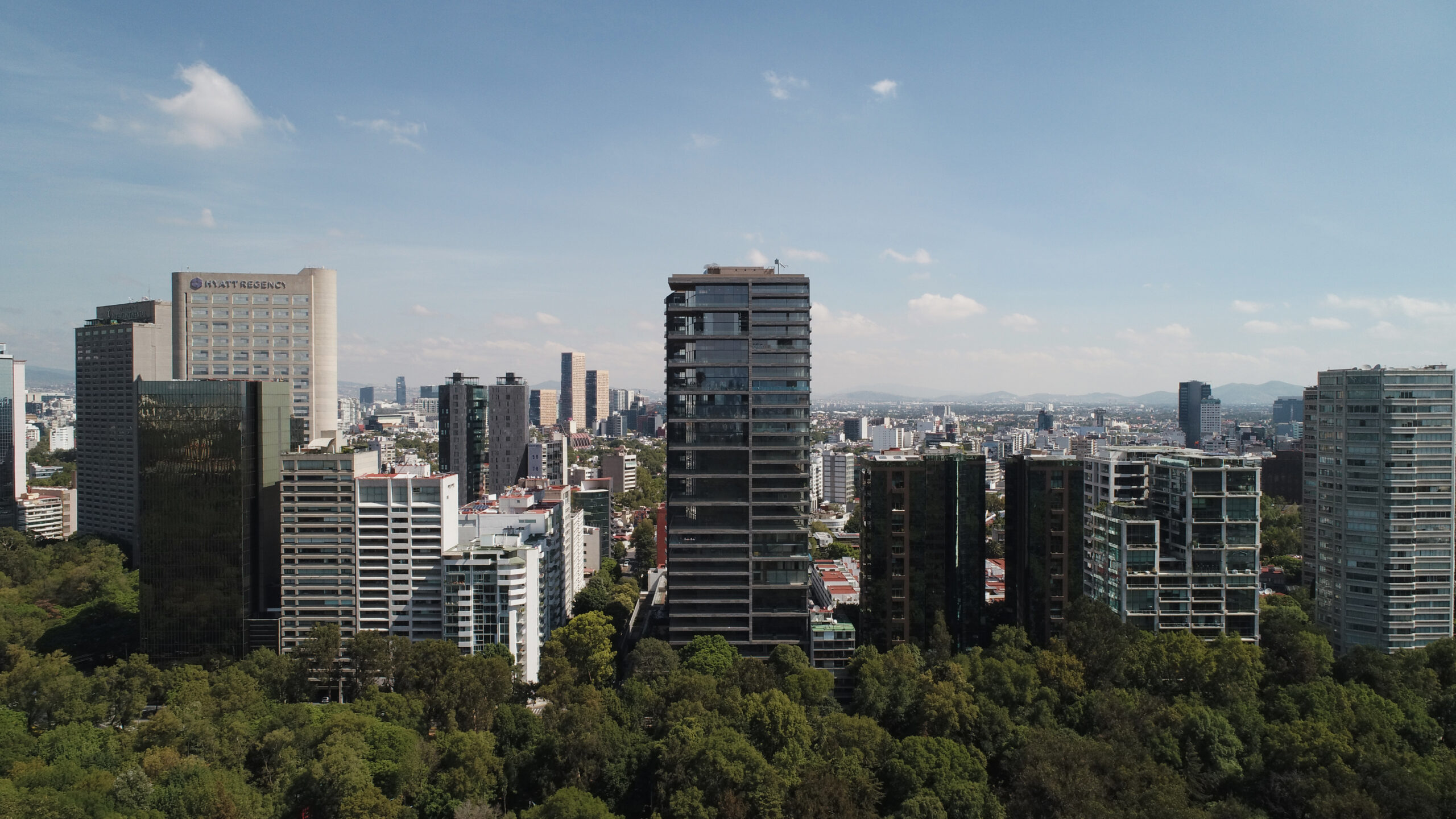 Daytime aerial photo of Rubén Darío 255 by Sordo Madaleno rising above the Bosque de Chapultepec tree canopy, with the Mexico City skyline and Hyatt Regency and InterContinental hotels in the background.