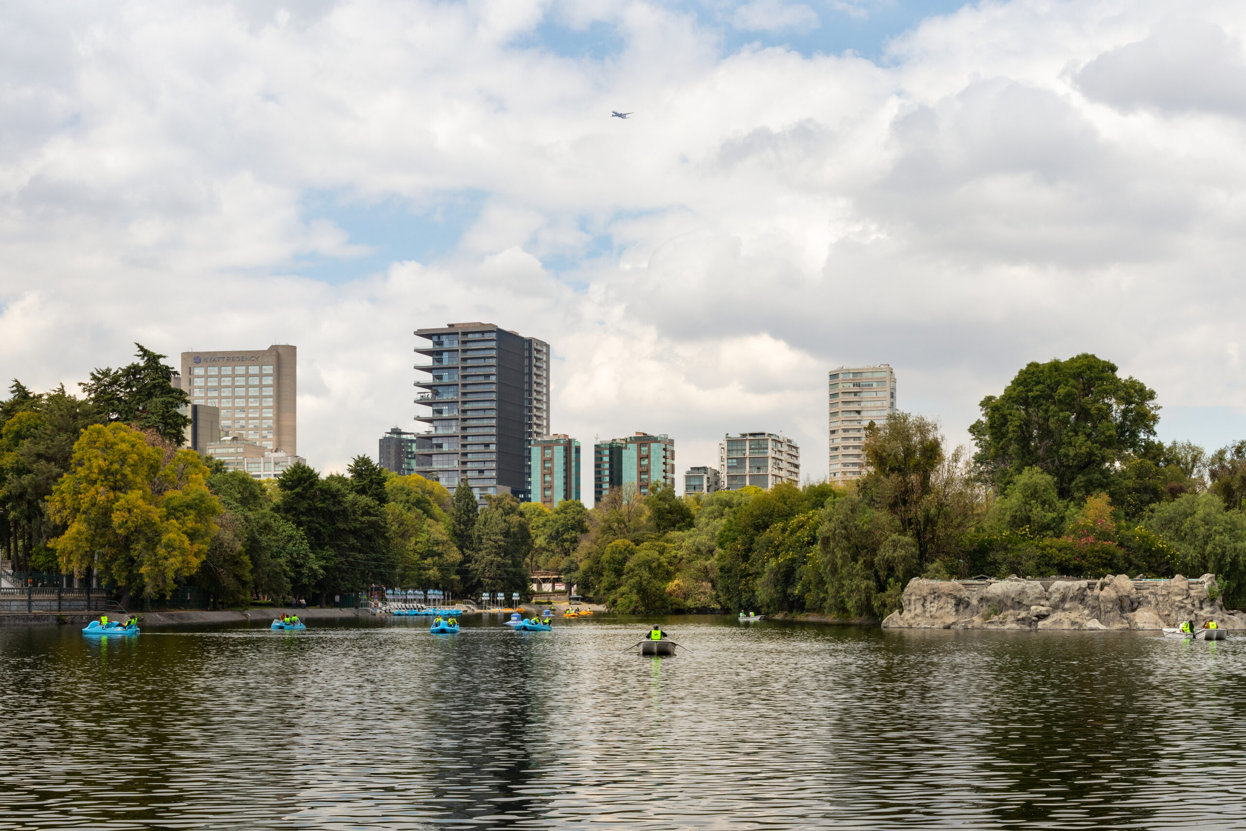 View of Rubén Darío 255 by Sordo Madaleno rising above the tree line beyond the boating lake of Bosque de Chapultepec, with paddle boats on the water.