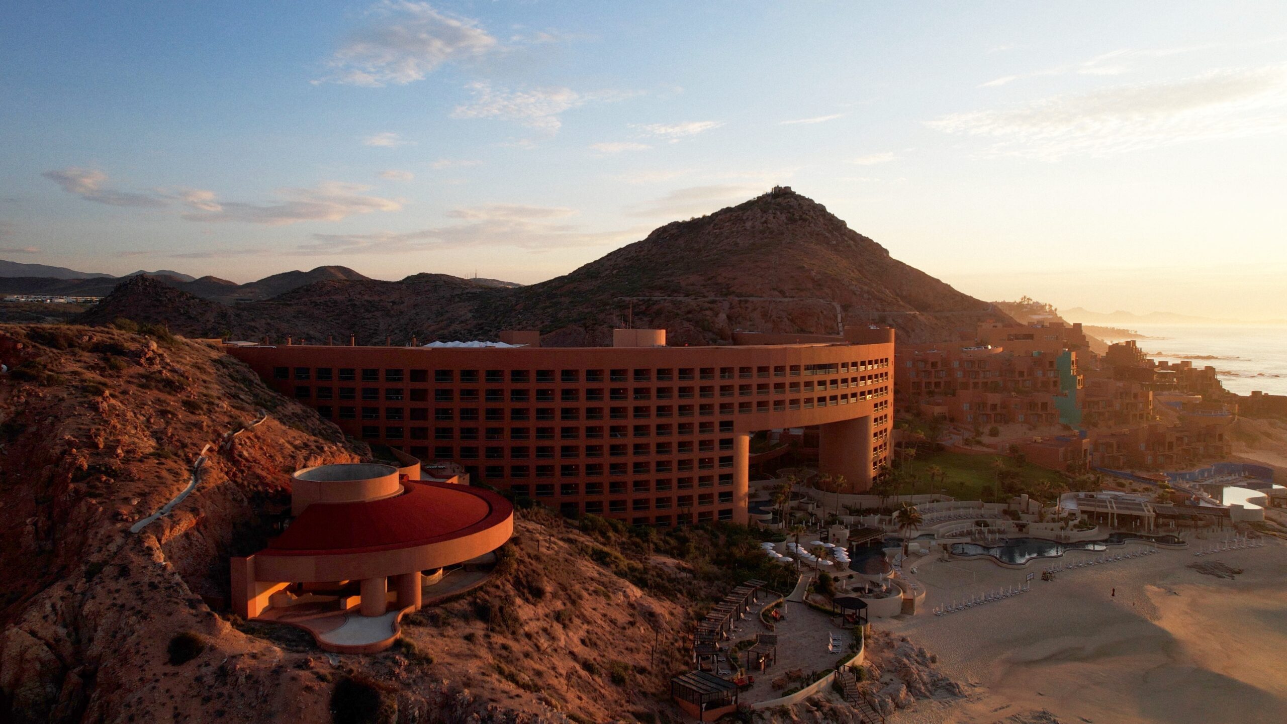 Aerial view of the Westin Regina Los Cabos hotel by Sordo Madaleno, its arched red volume spanning a rocky hillside at sunset